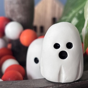 Peut inclure: Deux petites figurines de fantômes blanches, lisses, avec des sommets arrondis et des bases plates. Chaque fantôme présente deux yeux circulaires noirs et une bouche circulaire noire plus grande, créant une expression surprise.