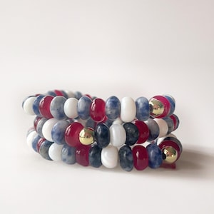 May include: Three beaded bracelets in red, white, blue, and gray. The bracelets are made of round beads and feature gold-colored accents. The bracelets are stacked on top of each other.