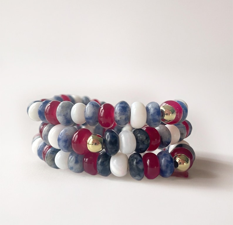 May include: Three beaded bracelets in red, white, blue, and gray. The bracelets are made of round beads and feature gold-colored accents. The bracelets are stacked on top of each other.