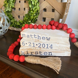 May include: A stack of four white wooden blocks with the names "matthew", "6.21.2014", and "kirsten" written in black. A strand of red wooden beads with a brown tassel is draped over the blocks.