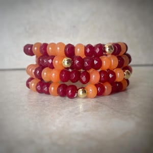 May include: A stack of four beaded bracelets. The bracelets alternate between orange and deep red faceted beads, with small gold-colored beads interspersed. The bracelets are stacked together, creating a layered effect.