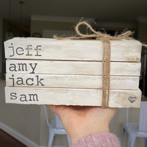 May include: A stack of four white painted books with the names "jeff", "amy", "jack", and "sam" printed on them. The books are tied together with twine and a small heart is on the bottom book.