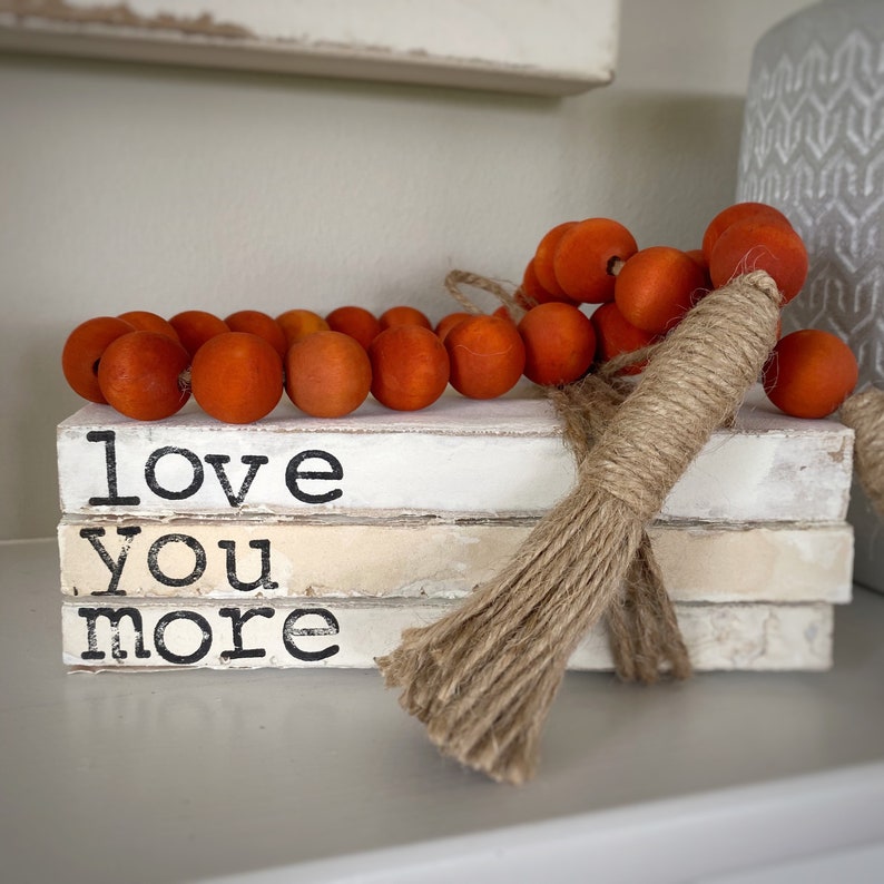 May include: A stack of white books with the words "love you more" printed on the front. The books are decorated with a garland of orange wooden beads and a brown jute tassel.
