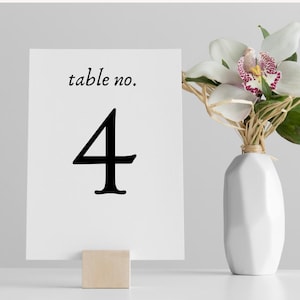May include: White card with black text that reads "table no. 4" on a wooden stand. The card is in front of a white vase with a white orchid.