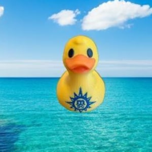 Op de afbeelding: Een gele badeend met een oranje snavel en een blauw embleem drijft in turquoise water. De achtergrond toont een helderblauwe lucht met witte wolken en een rotsachtige kustlijn.