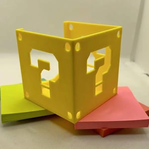 Puede incluir: Un bloque de interrogación amarillo impreso en 3D con un interior hueco. El bloque está sentado encima de tres notas adhesivas de colores.