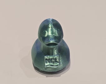 May include: A teal-colored duck figurine with a textured surface. The duck has a small beak and a rectangular emblem with the letters "NCL" on its chest. The figurine is set against a light background.
