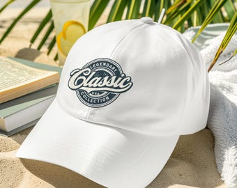 May include: White baseball cap with a navy blue embroidered logo that says "Legendary Classic USA Collection". The cap is on a sandy beach.