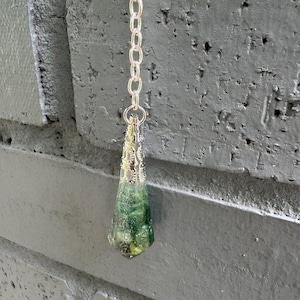 May include: A silver chain suspends a teardrop-shaped pendant. The pendant features a translucent green and yellow stone encased in a decorative silver filigree. The background is a gray brick wall.