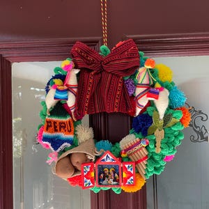 May include: A colourful Christmas wreath featuring a red bow, green pom-poms, and a nativity scene. The wreath also includes two white llama ornaments, a knitted bag with potatoes, and a sign that reads "PERU".