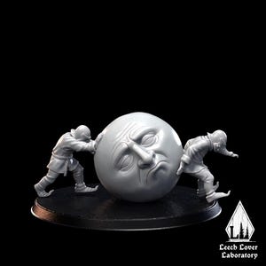 May include: A gray sculpture of two figures pushing a large, spherical moon with a face. The figures are dressed in medieval-style clothing. The moon has a sad expression. The sculpture is on a black base.