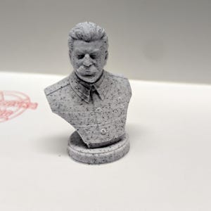 May include: A white 3D printed bust of a man with a serious expression. The bust is detailed with a textured surface and a patterned shirt.