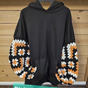Peut inclure: Sweat-shirt à capuche noir avec des manches en crochet orange, blanc et noir. "Digital Pattern Only" est écrit en bas de l'image.