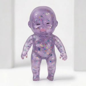 May include: A translucent purple baby doll with closed eyes and a slight smile. The doll is covered in colorful glitter, including stars and other shapes. The doll's fingernails are painted red.