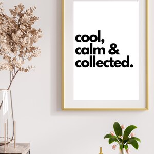 May include: A gold framed print with the text "cool, calm & collected." on a white background. The print is hanging on a white wall above a wooden table with a vase of flowers.