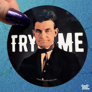 May include: A round black sticker featuring a portrait of a man in a suit with the words "TRY ME" in white. The sticker also includes the text "chaoticgoodtrouble.com" and "chaotic good trouble".