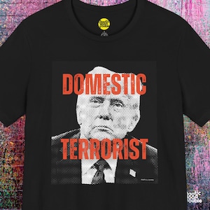 May include: Black t-shirt with a graphic of a man's portrait and the words "DOMESTIC TERRORIST" in red block letters. The shirt has a crew neck and short sleeves. The brand logo is visible on the upper right.