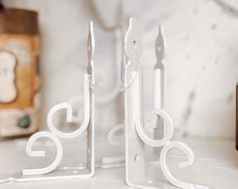 May include: White metal shelf brackets with decorative scrollwork and a stylized flame design. The brackets are designed to be mounted on a wall or other surface to support a shelf. The brackets are painted white.