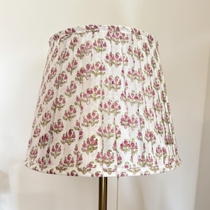 May include: A white lampshade with a pleated design and a repeating floral pattern in pink and green. The lamp has a brass-coloured metal base and a pull chain with a decorative ball.