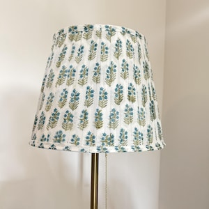 Peut inclure: Abat-jour plissé blanc orné d'un motif floral bleu et vert. Il est posé sur un pied de lampe en laiton. Une chaîne en laiton avec une petite boule pend de l'abat-jour.