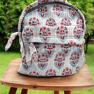 Puede incluir: Una mochila acolchada con un estampado floral en rosa, rojo y azul sobre un fondo gris claro. La mochila tiene un asa superior, un compartimento principal con cremallera y un bolsillo frontal. Está sobre un taburete de madera.