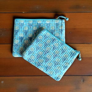 May include: Two quilted, zippered pouches in shades of blue with a floral pattern. The pouches have a light blue background with a repeating pattern of white and blue flowers. The zippers and pull tabs are a matching light blue.