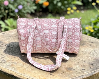 Könnte beinhalten: Eine rosa-weiß gemusterte Reisetasche mit zwei Griffen und einem Schultergurt. Die Tasche ist aus gestepptem Stoff gefertigt und steht auf einer Holzoberfläche. Der Hintergrund zeigt eine verschwommene Gartenszene.