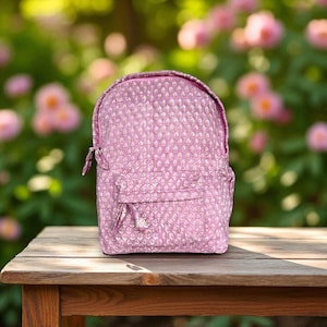 May include: A pink backpack with a white floral design. The bag has a front pocket, a top handle, and adjustable straps. It is on a wooden table, with a blurred floral background.