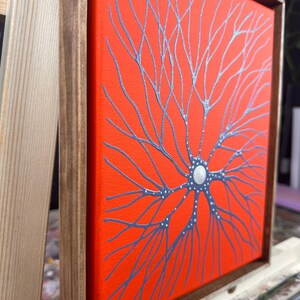 May include: A framed canvas painting with a bright orange background and a silver-grey tree-like design. The tree has a white circle in the center.