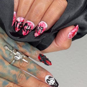 May include: Close-up of manicured nails with diverse designs. Some nails display a red and white marbled effect with black lettering. Other nails have black bead accents or a black and white graphic.