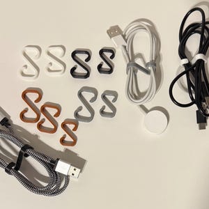 Puede incluir: Surtido de organizadores de cables en varios colores, incluyendo blanco, gris y marrón. Los organizadores tienen forma de letra 'S' y están diseñados para mantener los cables y los cables ordenados. También se incluyen dos cables de carga y una base de carga blanca.
