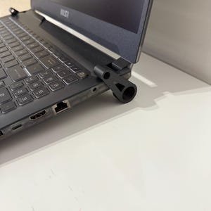May include: A black laptop computer with a black laptop stand attached to the hinge. The laptop is on a white surface.