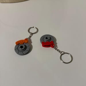 Peut inclure: Deux porte-clés avec des motifs de disques de frein miniatures gris et rouges. Les rotors ont des trous percés et sont associés à des étriers orange ou rouges. Chaque porte-clés comprend une chaîne argentée et un anneau fendu.