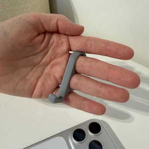 Op de afbeelding: Een grijze vingergrip voor een smartphone, ontworpen om om een vinger te lusssen voor een stevige grip. De grip is gemaakt van een flexibel materiaal en wordt getoond in gebruik met een zilveren smartphone.