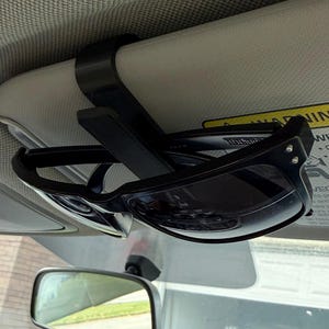 May include: A black plastic car visor clip holds a pair of black sunglasses. The sunglasses are folded in half and the clip is attached to the visor of a car.