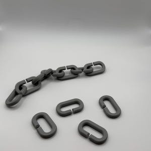Puede incluir: Una cadena gris con varios eslabones sueltos. La cadena está hecha de un material sólido y tiene un acabado mate. Los eslabones son de forma ovalada y tienen un diseño simple e industrial. El fondo es blanco liso.