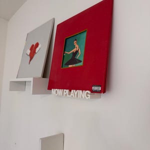 Puede incluir: Dos portadas de álbumes de vinilo montadas en una pared blanca. Una es roja con una mujer con un vestido negro, la otra es gris con un corazón rojo. Un letrero blanco "NOW PLAYING" está debajo del álbum rojo.