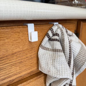 May include: Two white plastic hooks mounted under a cabinet, with a brown and white striped kitchen towel hanging from one of the hooks.