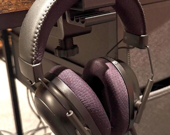 May include: Black headphones with a black clamp-on desk stand. The headphones have a padded headband and earcups.
