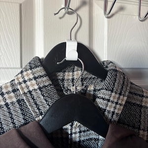 May include: Two black wooden clothes hangers with garments hanging on a chrome over-the-door hook. The top garment is a black and white plaid jacket. The bottom garment is a brown jacket. The hangers are attached to a white door.