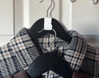 May include: Two black wooden clothes hangers with garments hanging on a chrome over-the-door hook. The top garment is a black and white plaid jacket. The bottom garment is a brown jacket. The hangers are attached to a white door.