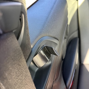 May include: A close-up of a black car door handle with a silver metal accent. A grey seatbelt is visible on the left. The door panel is black and has a textured surface.