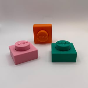 May include: Three toy bricks in pink, orange, and green. Each brick has a raised circular top with the word "BRICK" embossed on it. The bricks are square-shaped and sit on a white surface.