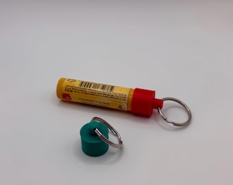 Könnte beinhalten: Eine gelb-rote Signalrakete mit einem silbernen Schlüsselring neben einem türkisgrünen Stopfen mit einem silbernen Schlüsselring. Die Rakete hat ein gelbes Etikett mit schwarzem Text und einem Barcode. Der Stopfen ist zylindrisch.