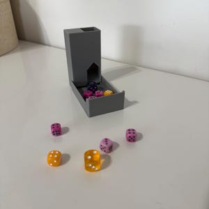May include: A grey dice tower with an open tray, surrounded by scattered dice. The dice are various colours, including pink, yellow, and clear orange. Each die has white dots. The tower is on a white surface.