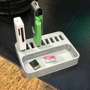 Könnte beinhalten: Grauer Plastik-Organizer mit mehreren Steckplätzen zum Verstauen von USB-Sticks, Speicherkarten und anderen kleinen elektronischen Geräten. Der Organizer hat ein kleines Fach zum Aufbewahren von zusätzlichen Gegenständen. Ein grüner USB-Stick, ein weißer USB-Stick und eine rosa Speicherkarte sind im Organizer sichtbar.
