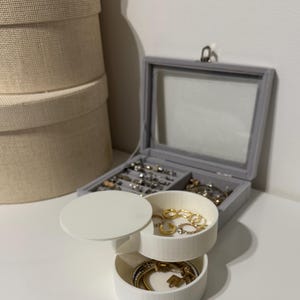 Op de afbeelding: Een grijze sieradendoos met een glazen deksel en meerdere compartimenten, gevuld met diverse ringen, kettingen en armbanden. Een witte, gelaagde sieradenorganizer is open en toont gouden en zilveren sieraden. Gestapelde beige hoeden dozen staan op de achtergrond.