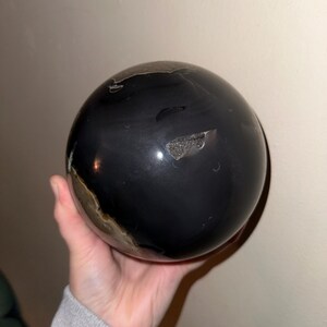Puede incluir: Una esfera negra pulida con una pequeña sección marrón claro. La esfera está hecha de piedra natural y tiene un acabado liso y brillante.