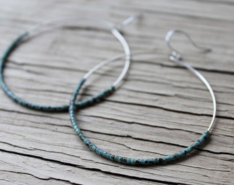 May include: A pair of hoop earrings with a silver-colored metal frame. The lower portion of each earring is adorned with small, turquoise-colored beads. The earrings are displayed on a weathered wooden surface.