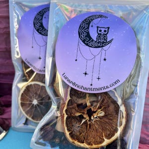 May include: Two clear plastic bags filled with dried fruit, herbs, and spices. The bags have a purple and black sticker with a crescent moon and an owl on it. The text on the sticker reads "Lunar&Enchantments.com".